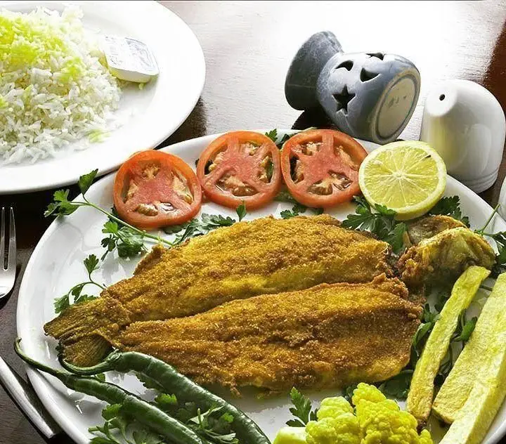 ماهی سرخ شده طلایی رستوران ایرانی محفل همراه با برنج، لیمو، گوجه فرنگی، فلفل سبز و سبزیجات تازه. غذایی لذیذ و اصیل ایرانی.
