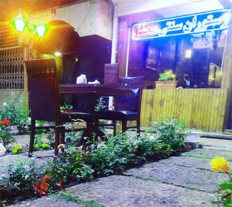 فضای باز دلنشین رستوران ایرانی محفل در شب، با میز و صندلی‌های چوبی شیک و باغچه‌های پر از گل، محیطی ایده‌آل برای صرف شام.