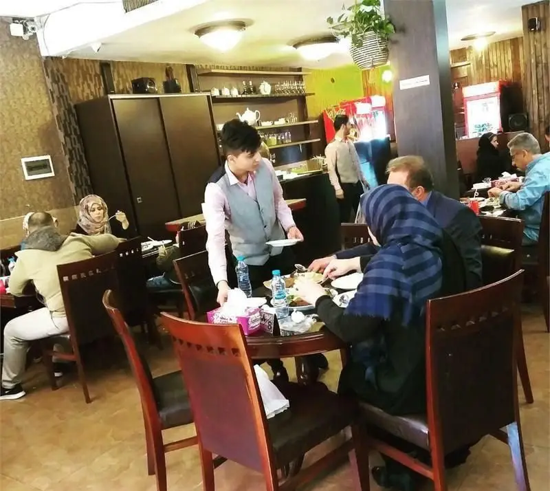 محیط داخلی رستوران ایرانی محفل، با حضور مشتریان در حال صرف غذا و پذیرایی یک گارسون. فضایی دلنشین برای وعده‌های خانوادگی.