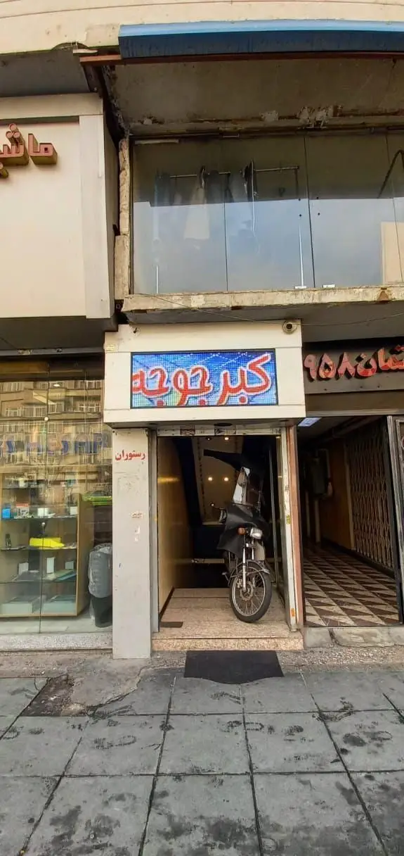 تصویری از نمای بیرونی رستوران اکبر جوجه با تابلوی LED روشن و ورودی اصلی آن که یک موتور سیکلت پارک شده است.