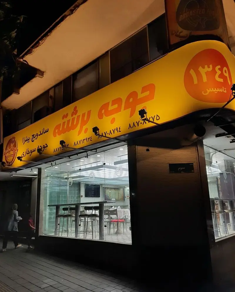 نمای شبانه فست فود جوجه برشته، با تابلوی روشن خدمات ساندویچ، پیتزا و جوجه سوخاری، رستورانی مدرن تاسیس ۱۳۶۹.
