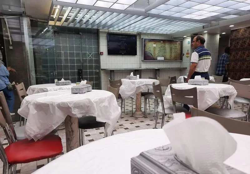 تصویر فضای داخلی فست فود جوجه برشته با میزهای گرد، صندلی‌های راحت و رومیزی‌های یکبار مصرف سفید، منوهای غذا روی دیوار قابل مشاهده است.