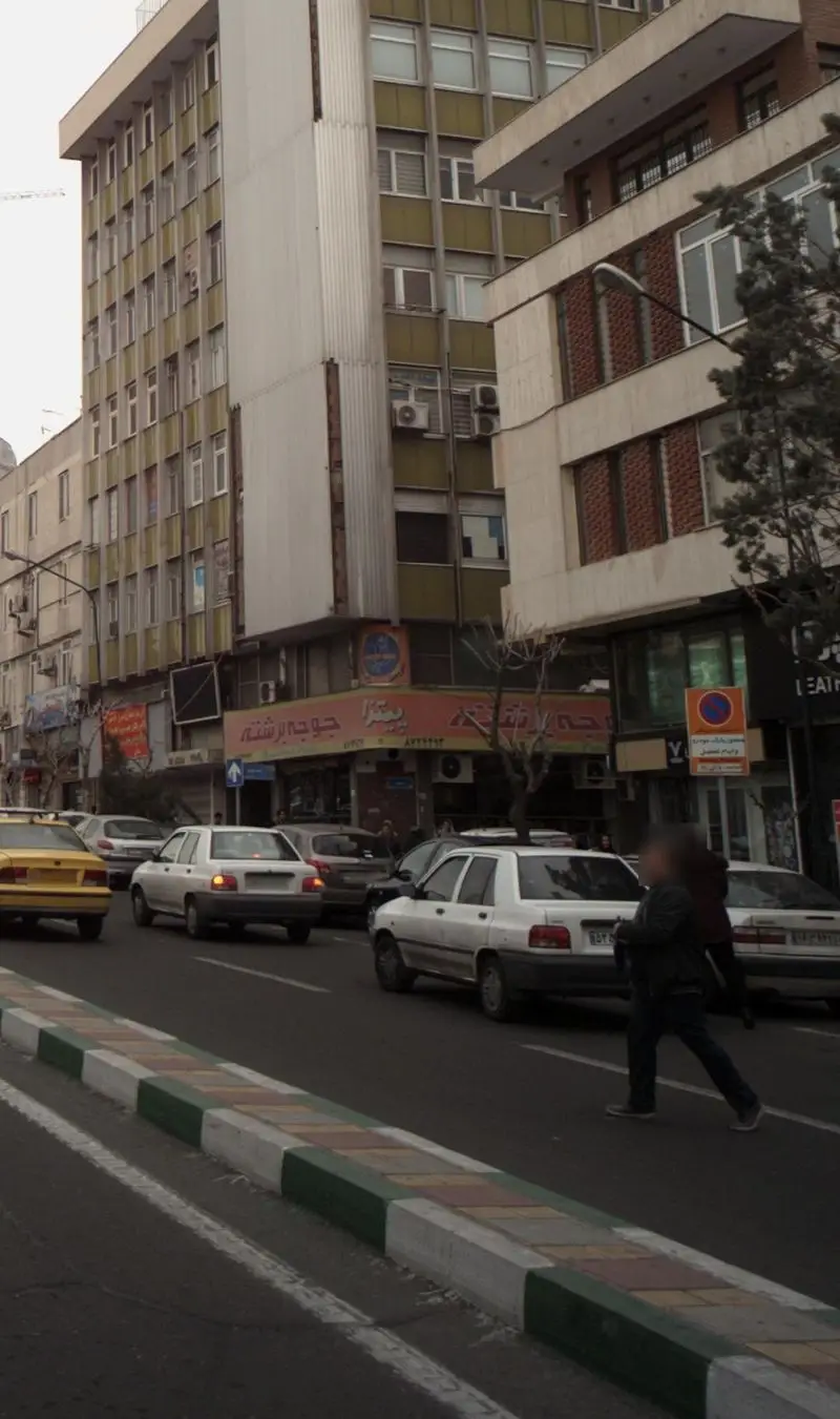 نمای بیرونی فست فود جوجه برشته در خیابانی شلوغ و پر رفت و آمد شهری، همراه با خودروهای متعدد و عابران پیاده.