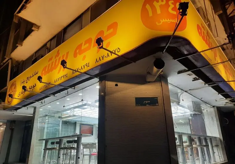 فست فود جوجه برشته: نمای شبانه با تابلو روشن زرد، منوی ساندویچ، پیتزا و برگر به همراه شماره تماس. واقع در خیابان سوم تهران.