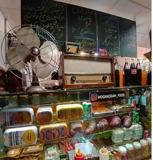 داخل ساندویچی مقدم: ویترین متنوع کالباس، سوسیس، غذاهای آماده، نوشیدنی‌ها. منوی تخته‌ای و دکور نوستالژیک با فن و رادیو قدیمی.