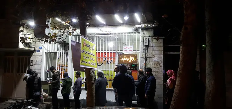 تصویری در شب از مشتریانی که برای خرید فلافل کیلویی میخکوب صف کشیده‌اند. مغازه با نورپردازی روشن، نمایی از کسب و کار پررونق.