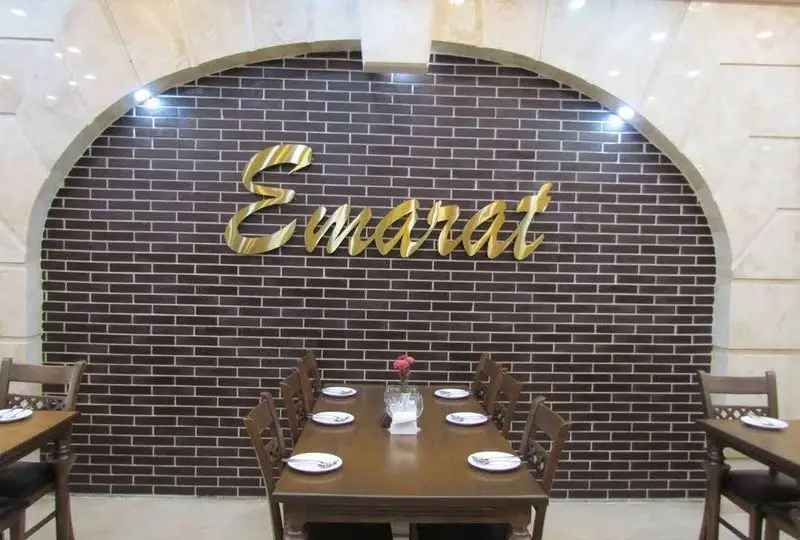 نمایی از دکوراسیون داخلی رستوران عمارت با دیوار آجری قهوه‌ای، تابلوی طلایی Emarat و میز و صندلی‌های چوبی برای مشتریان.