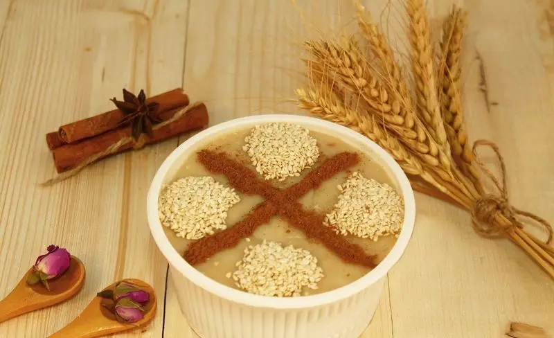 تصویری زیبا از حلیم گندم سنتی و خوشمزه دارچین، تزئین شده با کنجد و پودر دارچین، در کنار چوب دارچین و گندم برای صبحانه.