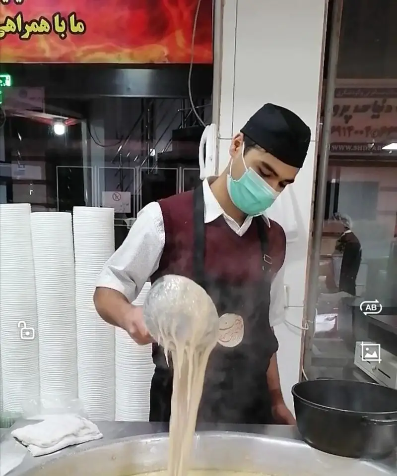 آشپز آش و حلیم دارچین ماسک‌زده، مشغول آماده‌سازی و سرو حلیم داغ و لذیذ در محیط بهداشتی با کاسه‌های یکبار مصرف.