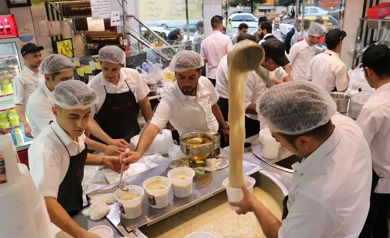 پرسنل مجرب آش و حلیم دارچین در حال پخت و سرو آش و حلیم سنتی ایرانی. محیط شلوغ آشپزخانه با کارمندان جوان و فعال، آماده‌سازی غذا برای مشتریان.