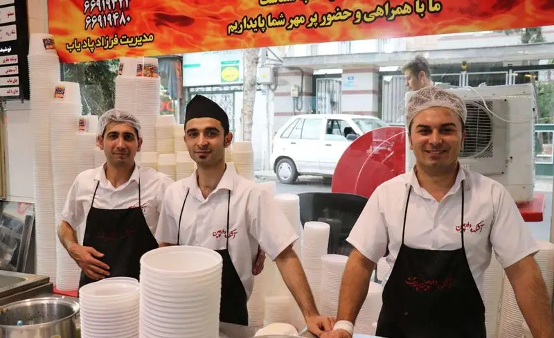 سه کارمند خوش‌رو در آش و حلیم دارچین، با مدیریت فرزاد پادیاپ، پشت پیشخوان و ظروف یکبار مصرف، آماده سرو غذای گرم ایرانی.