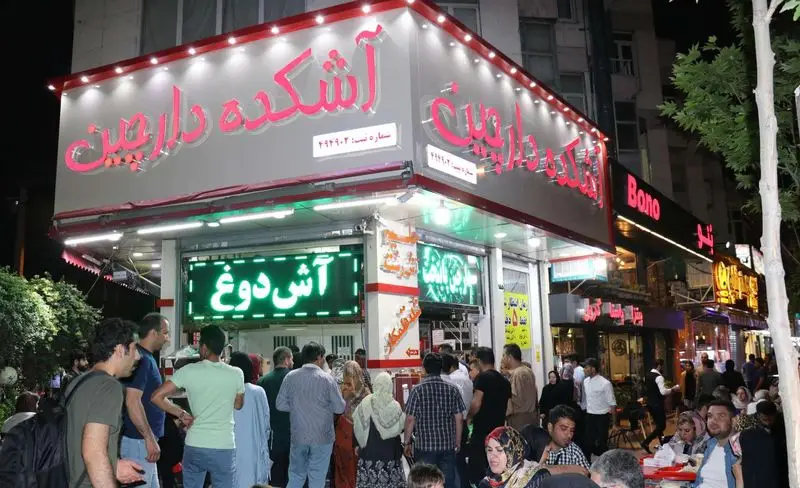 نمای بیرونی آش و حلیم دارچین در شبی پر جنب و جوش، با تابلوی روشن آش دوغ و مشتریان زیادی در حال لذت بردن از غذای سنتی ایرانی.