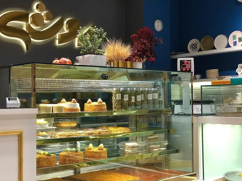 تصویری از ویترین شیشه‌ای شیرینی لبنانی روشه پر از کیک‌ها، دسرها و شیرینی‌های تازه و خوشمزه. دکوراسیون داخلی شیک و مدرن.