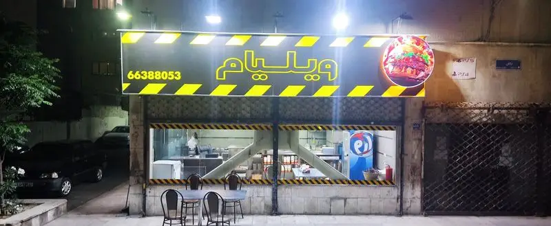 فست فود ویلیام، نمای شب از مغازه با تابلوی روشن، شماره تماس ۶۶۳۸۸۰۵۳ و تصویر همبرگر. میز و صندلی بیرون آماده پذیرایی است.