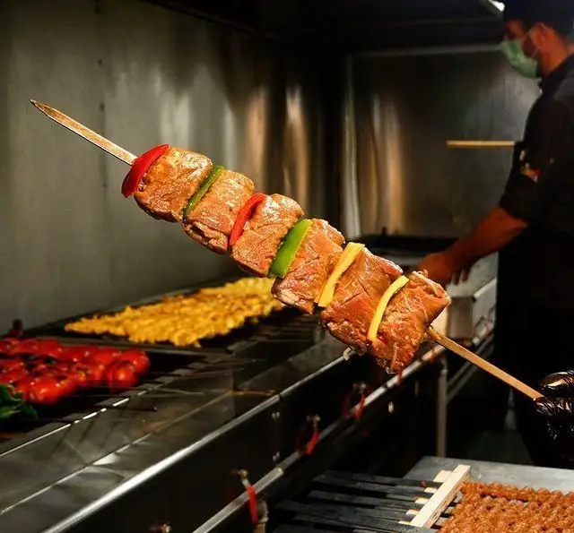 نمایی از آشپز رستوران بین المللی ۱۳۳ در حال آماده سازی سیخ کباب گوشت و فلفل دلمه‌ای رنگارنگ روی گریل، با کباب کوبیده آماده پخت.