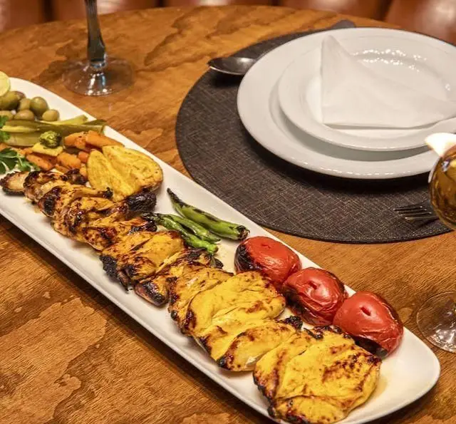 جوجه کباب لذیذ و اصیل ایرانی رستوران بین المللی ۱۳۳، سرو شده با گوجه و فلفل کبابی، ترشی و دورچین اشتهاآور روی میز چوبی