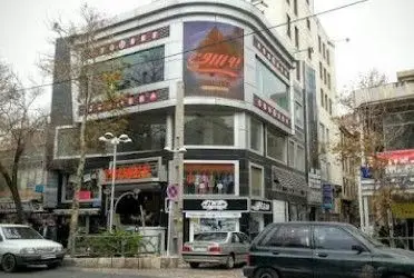 نمای بیرونی ساختمان تجاری مدرن و شیک در تهران، شامل کافه دیاکو و فروشگاه‌های متنوع، با ماشین‌ها و فضای شهری پویا در خیابان.