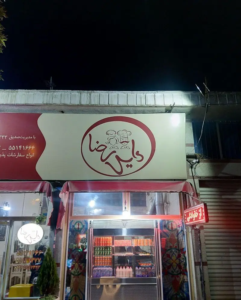 نمای شب رستوران دایی رضا در مشهد، با تابلوی آشپزها. عرضه کننده فست فود، فالوده و انواع غذاها. پذیرش سفارشات تلفنی.