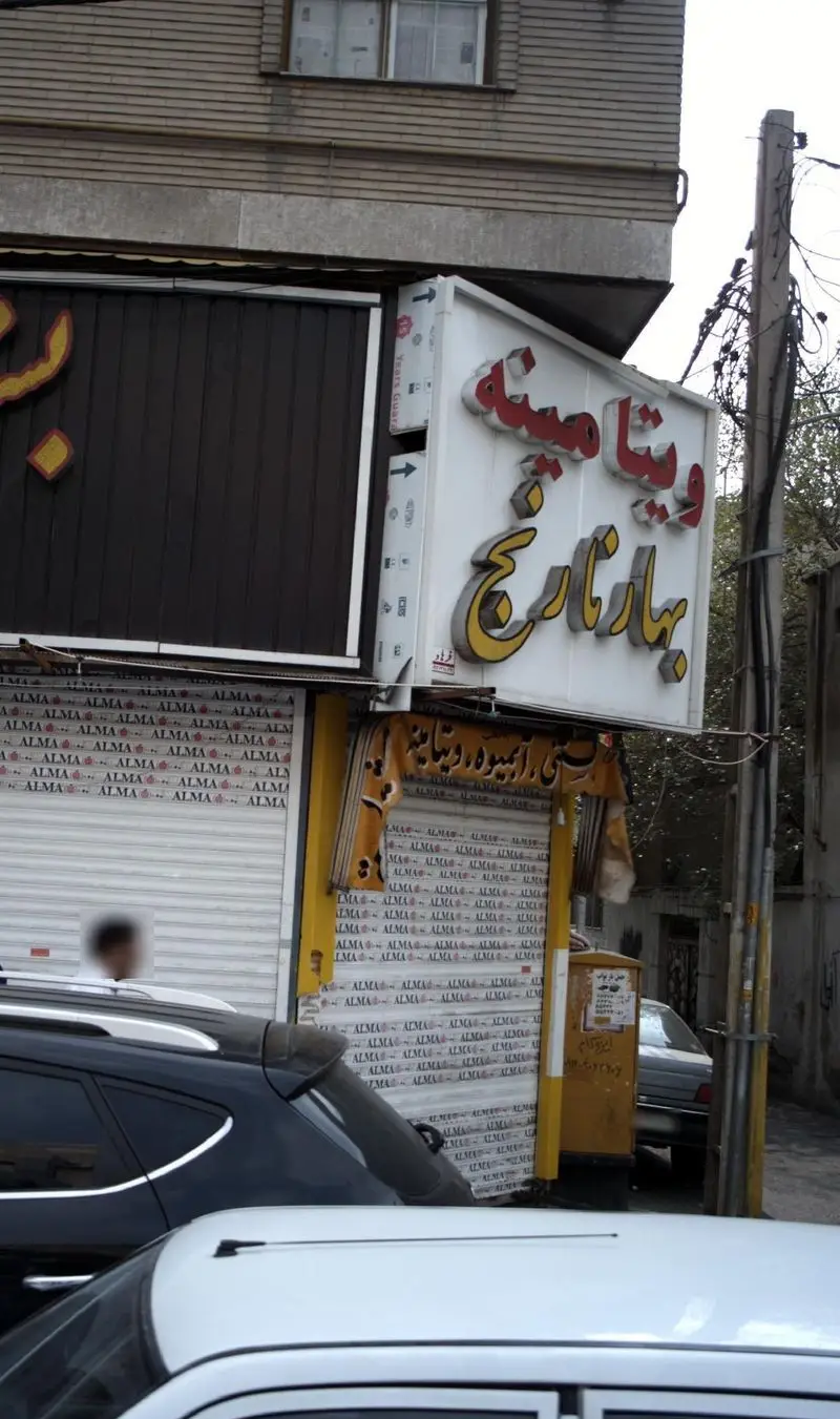 تصویر نمای بیرونی مغازه ویتامینه بهار نارنج با تابلوی بزرگ قرمز و زرد، کرکره‌های بسته و خودروهای پارک شده در روز.