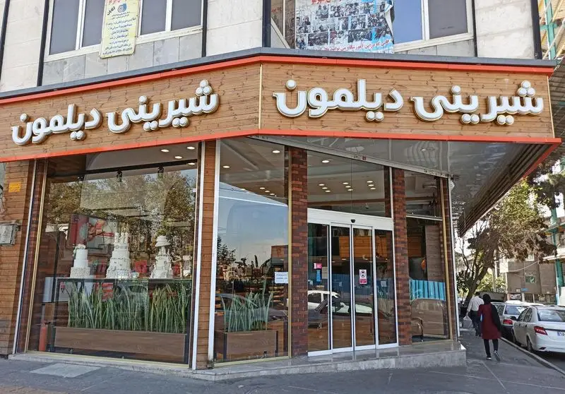 نمای جلوی قنادی شیرینی دیلمون، فروشگاهی مدرن با تابلوی روشن، ویترین شیشه‌ای بزرگ پر از کیک و شیرینی‌های متنوع و ورودی مهمان‌نواز.