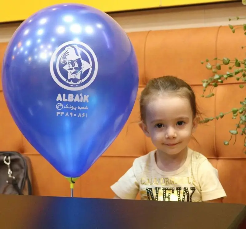 دختربچه‌ای شاد در رستوران البیک شعبه پونک با بادکنک آبی لوگودار. تجربه‌ای خانوادگی و غذای کودک لذت‌بخش در محیطی دوستانه.