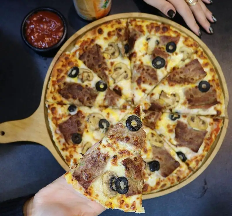 پیتزا گوشت و قارچ رستوران البیک با پنیر طلایی، زیتون سیاه و سس مخصوص، روی تخته سرو چوبی، یک برش در دست.