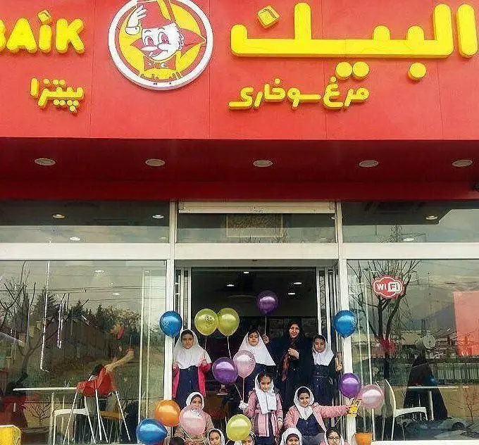 رستوران فست فود البیک با تابلوی روشن، شامل مرغ سوخاری و پیتزا، و جمعی از خانم‌ها و دختران با بادکنک در مقابل درب.