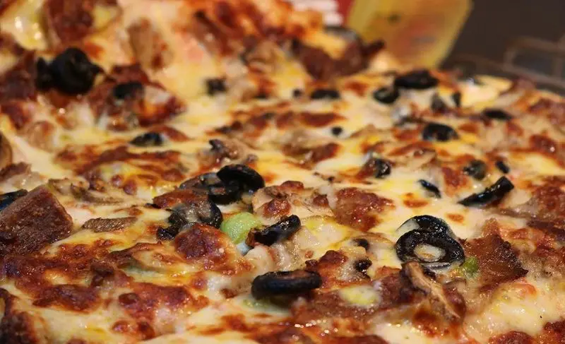 پیتزای داغ و لذیذ رستوران البیک با رویه فراوان پنیر طلایی، زیتون سیاه، قارچ و تکه‌های گوشت، آماده سرو در محیطی دلپذیر.