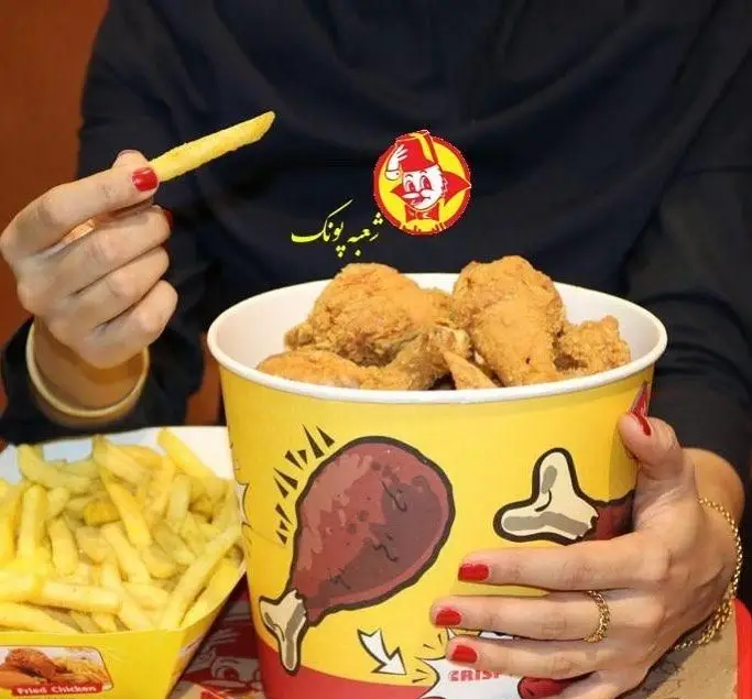 سطل پر از مرغ سوخاری ترد رستوران البیک در کنار سیب‌زمینی سرخ‌کرده طلایی، همراه با دستی که یک سیب‌زمینی را می‌گیرد. طعم لذیذ.