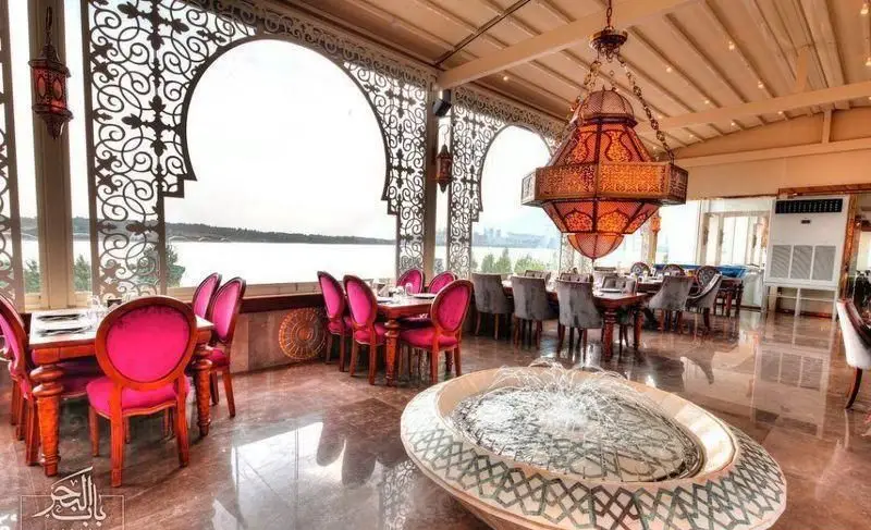 فضای داخلی رستوران لبنانی باب البحر با دکوراسیون سنتی عربی، میزهای چوبی و صندلی‌های شیک صورتی و خاکستری، مشرف به منظره دریاچه و آب‌نما.