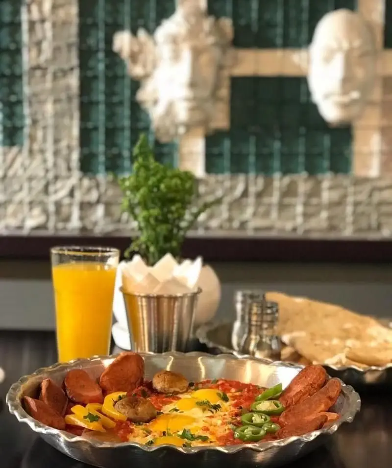 میز صبحانه لذیذ در کافی شاپ پرسوناژ: املت با سوسیس و مخلفات، نان تازه و آب پرتقال. فضایی دلنشین برای شروع روز.