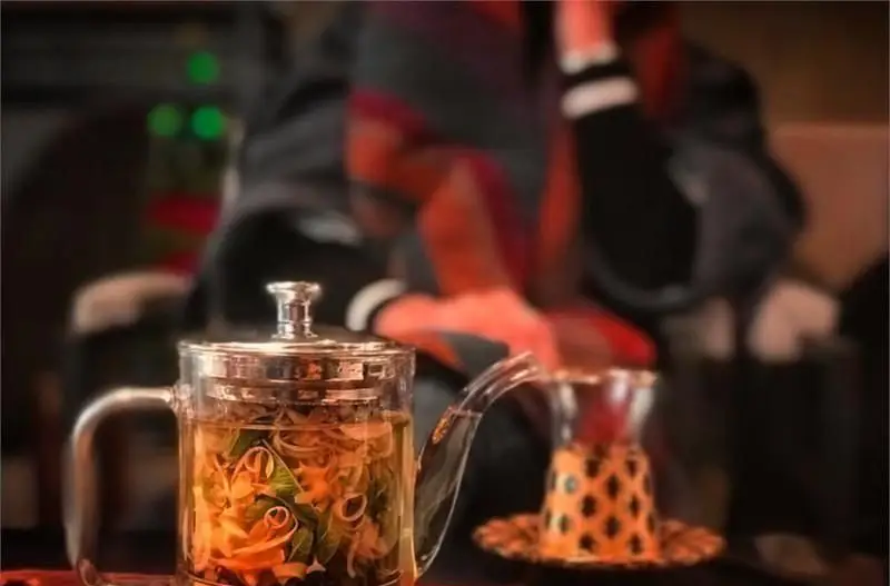 قوری دمنوش گیاهی شفاف در کافی شاپ پرسوناژ، نمایشگر نوشیدنی آرامش‌بخش با برگ نعناع و پوست پرتقال در فضایی دلنشین.