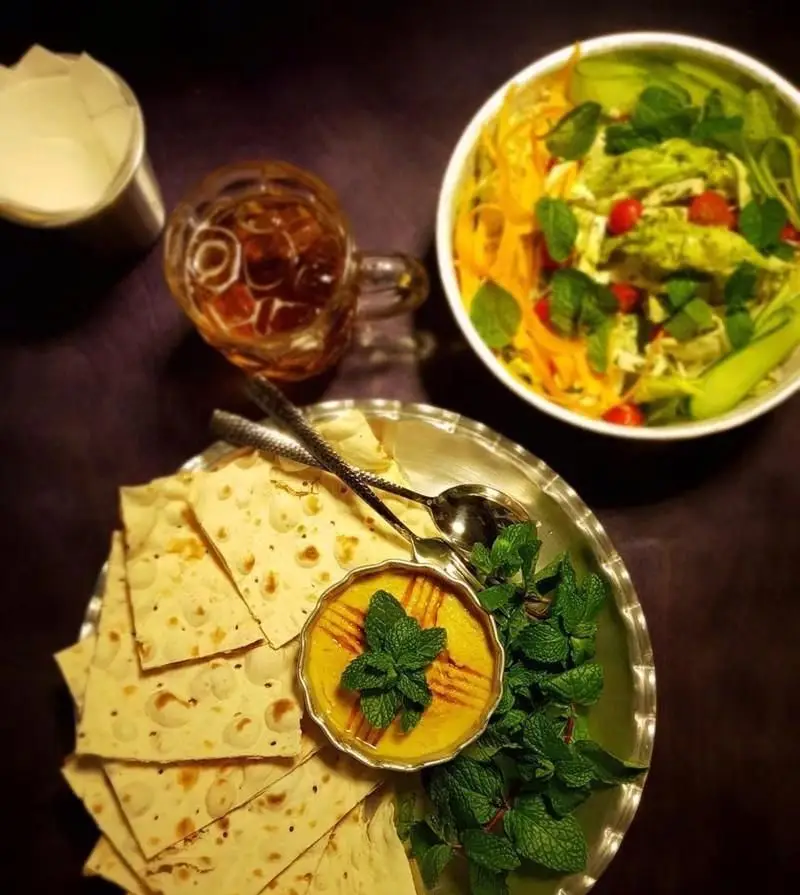 تصویری دلنشین از میزی در کافی شاپ پرسوناژ: سالاد تازه و رنگارنگ، حمص لذیذ با نان ترد و نعنای تازه، و نوشیدنی گوارا.