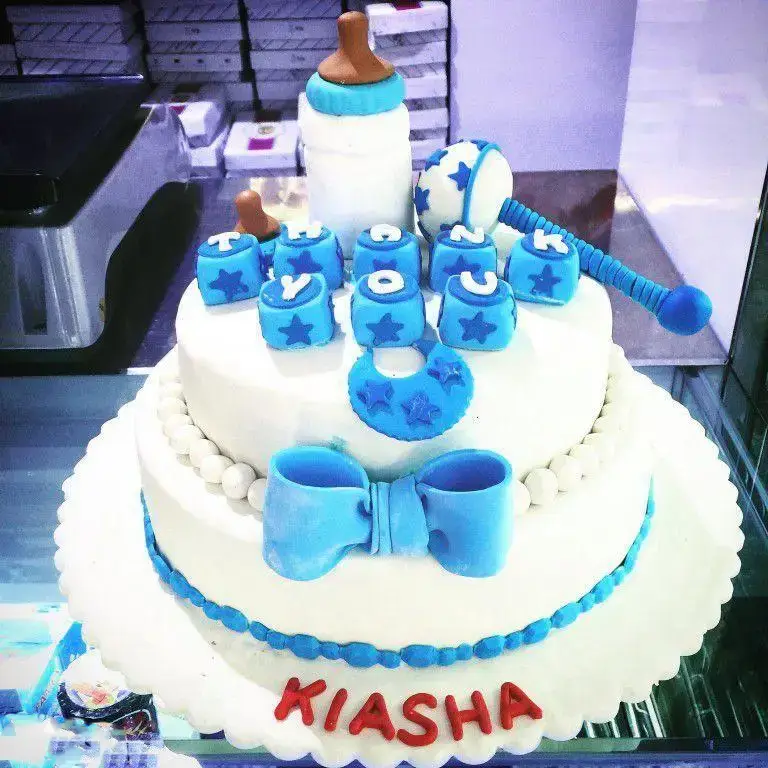 کیک تولد کیاشا، دو طبقه سفید با تزیینات آبی بچه‌گانه (شیشه شیر، جغجغه، بلوک حروف و ستاره) و مروارید از شیرینی لادن طلایی.