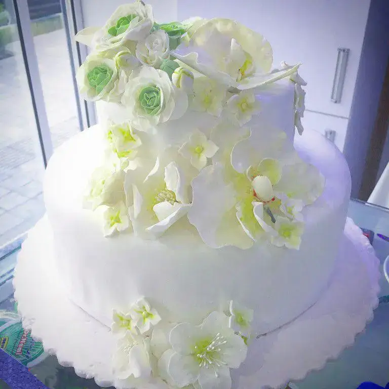 کیک عروسی سفید دو طبقه مجلل با تزیینات ظریف گل‌های شکری ارکیده و رز سبز و سفید. اثری هنری از شیرینی لادن طلایی.