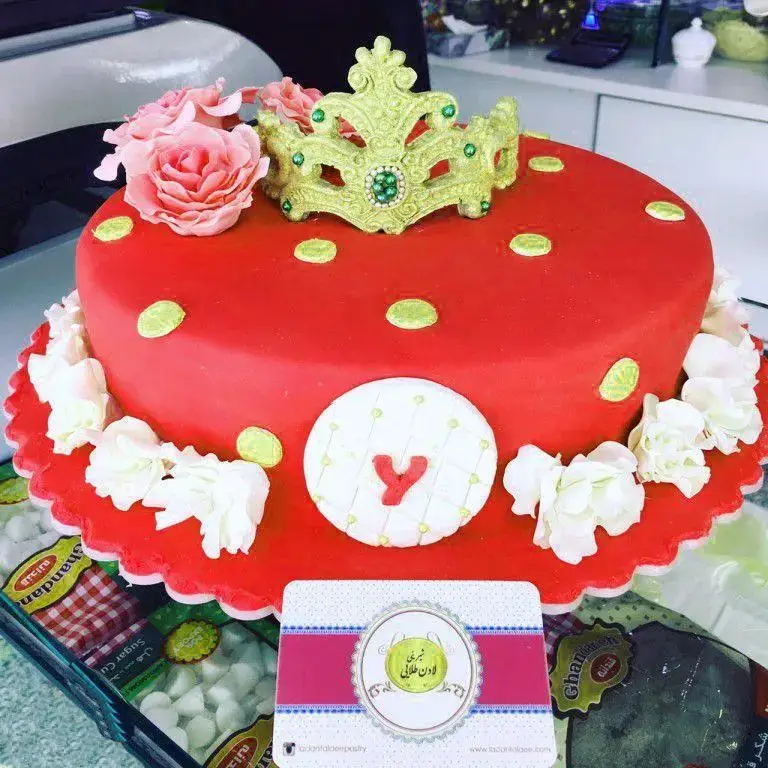 کیک تولد قرمز خیره‌کننده با تزئینات تاج طلایی، گل رز صورتی و پولکا دات سبز از شیرینی لادن طلایی. ایده‌آل برای جشن‌ها.