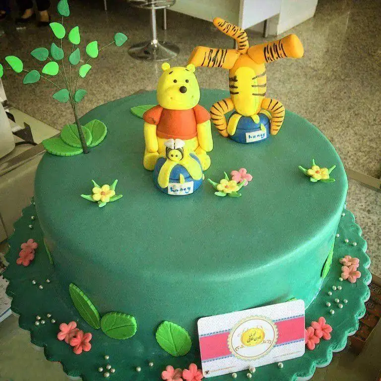 کیک تولد تم وینی پو و تیگر از شیرینی لادن طلایی، با تزئینات فوندانت، برگ، گل و ظرف عسل، مناسب جشن کودک.