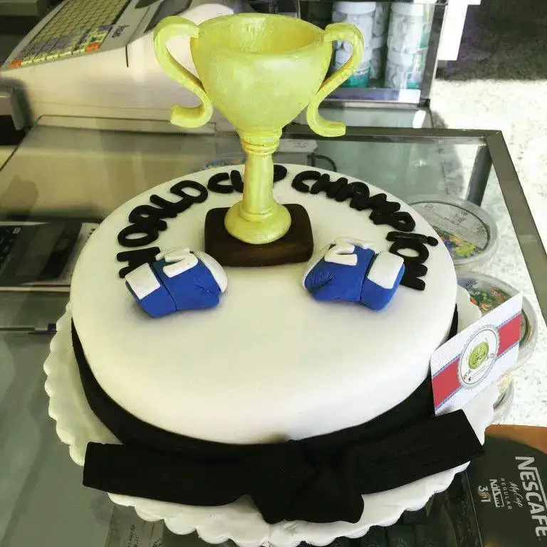 کیک سفارشی تم قهرمانی ورزشی شیرینی لادن طلایی شامل کاپ طلایی، دستکش بوکس و کمربند مشکی، مناسب جشن افتخارات و پیروزی.