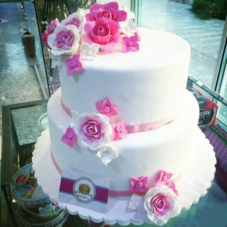 کیک دو طبقه سفید با تزیین گل‌های شکری صورتی و سفید و روبان‌های صورتی، اثری زیبا از شیرینی لادن طلایی برای مراسم خاص شما.
