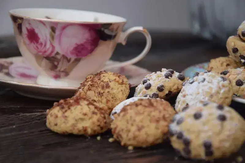 کوکی های متنوع شیرینی لادن طلایی با چیپس شکلات و رویه مغزدار، در کنار فنجان چای گلدار برای یک عصرانه دلپذیر.