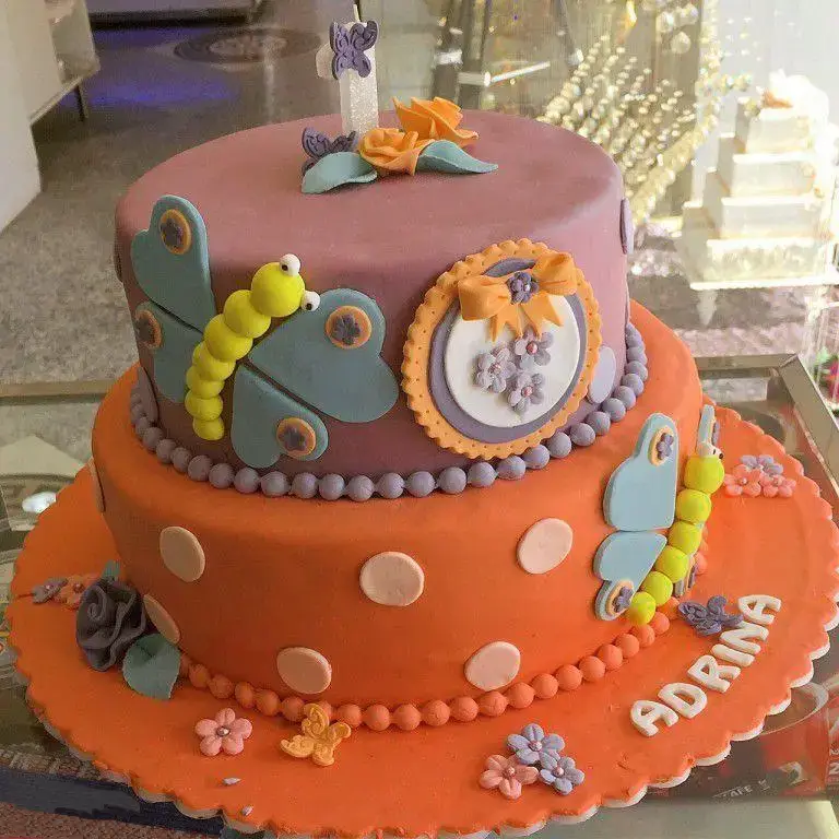 کیک تولد دخترانه دو طبقه با تم پروانه، رنگ‌های نارنجی و بنفش، تزئینات کاترپیلار و گل، مناسب جشن تولد یکسالگی آدرینا از شیرینی لادن طلایی.