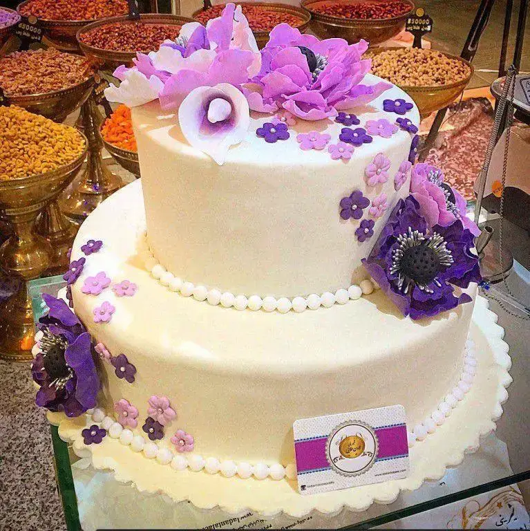 کیک تولد دو طبقه سفید شیرینی لادن طلایی با تزیینات گل‌های شکری بنفش و صورتی و مروارید. مناسب جشن‌ها و مراسم خاص.