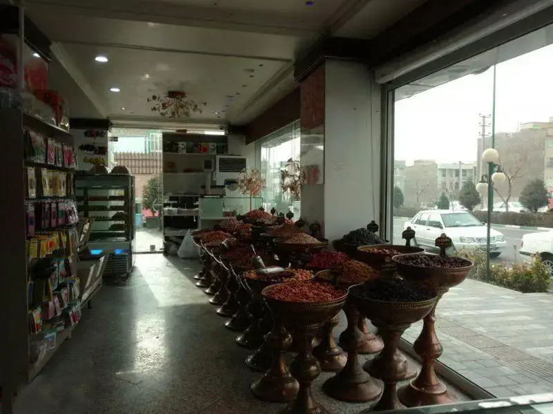 تصویری از داخل فروشگاه شیرینی لادن طلایی، شامل ردیفی از ظروف مسی پر از انواع آجیل و خشکبار، در کنار ویترین شیرینی‌جات و محصولات متنوع دیگر.