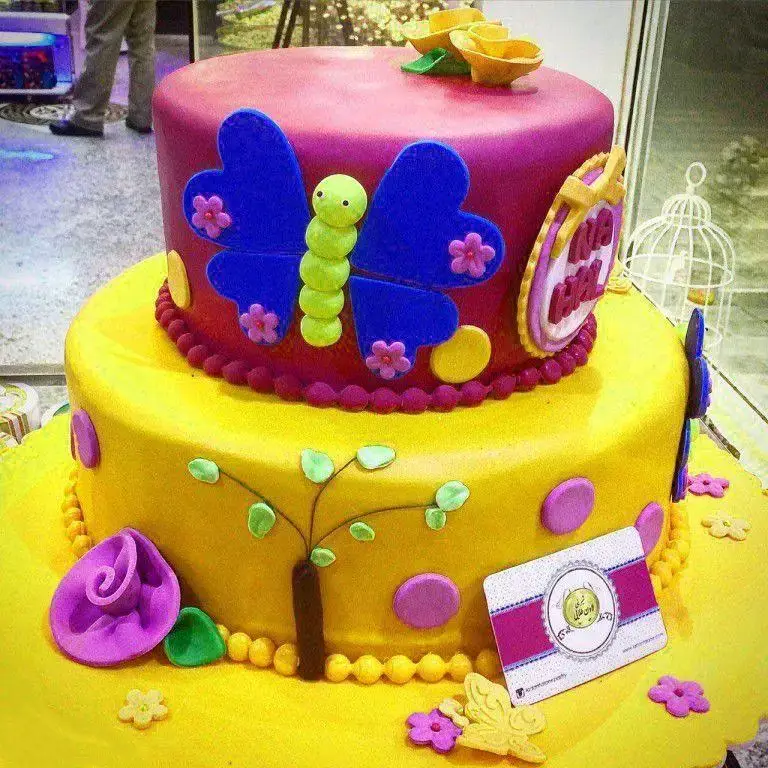 کیک تولد دو طبقه فانتزی شیرینی لادن طلایی با تزیین پروانه، کاترپیلار و گل‌های رنگارنگ، انتخابی ایده‌آل برای جشن‌ها.