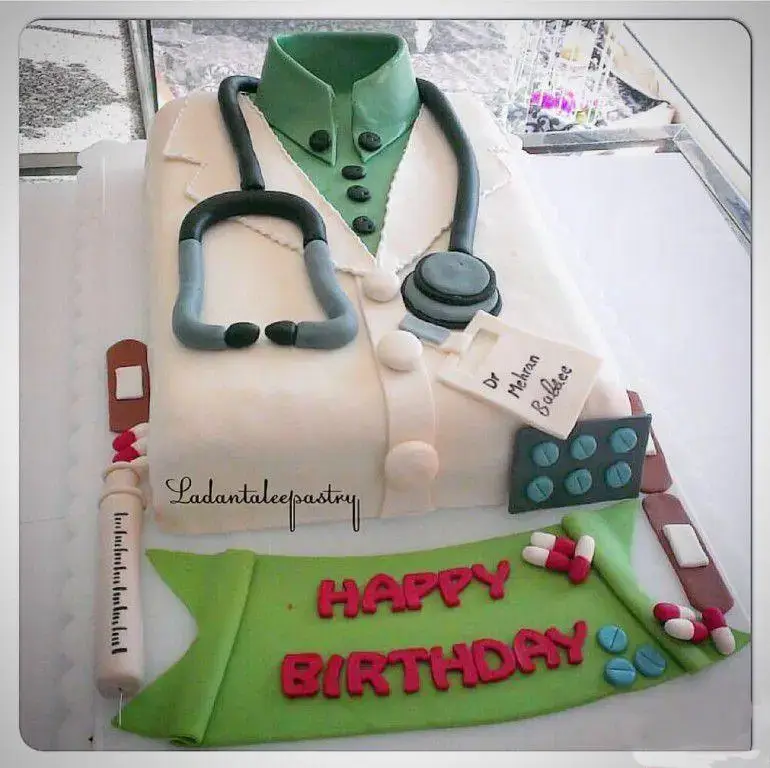 کیک تولد خاص به شکل روپوش دکتر با گوشی پزشکی، سرنگ، قرص و نوشته Happy Birthday، کاری از شیرینی لادن طلایی.