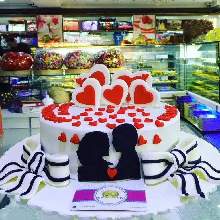 کیک سفید شگفت‌انگیز و عاشقانه با تزیین قلب‌های قرمز و طرح نیم‌رخ زوج، محصولی از شیرینی لادن طلایی. ایده‌آل برای سالگرد و ولنتاین در بهترین شیرینی فروشی.