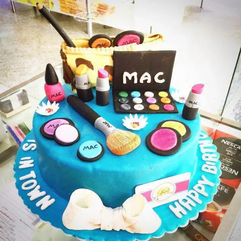 کیک تولد دخترانه آبی رنگ با تزیین لوازم آرایشی معروف مک (MAC) مانند رژلب و سایه چشم، آماده در شیرینی لادن طلایی.