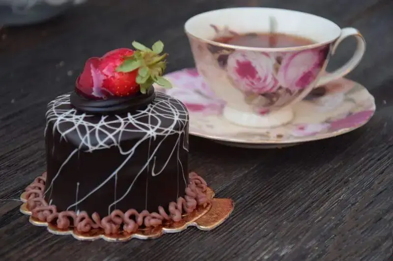 شیرینی لادن طلایی: کیک شکلاتی کوچک با تزئین توت فرنگی و چای در فنجان گل‌دار. دسری دلپذیر برای لحظات شیرین شما.