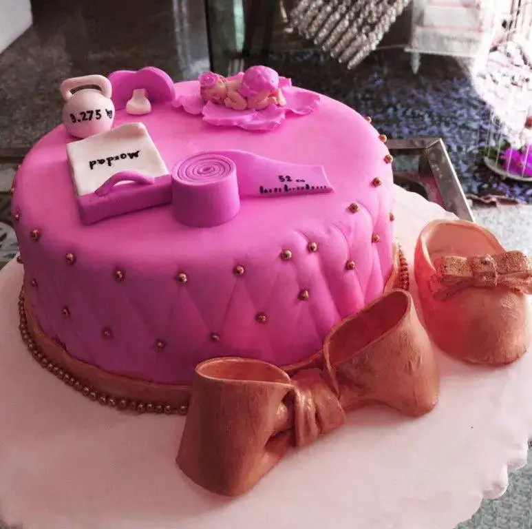 کیک سفارشی صورتی با تم نوزاد و تناسب اندام، شامل تزیینات وزنه، متر، مجسمه نوزاد و کفش نوزادی، از شیرینی لادن طلایی.