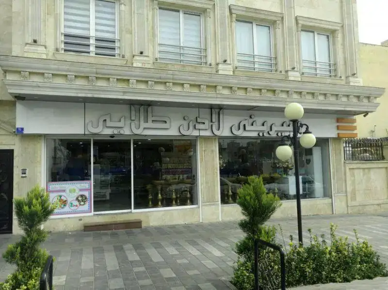 نمای بیرونی و تابلو بزرگ فروشگاه شیرینی لادن طلایی، با ویترین‌های پر از محصولات و پیاده‌رو تمیز در جلو. مناسب برای جستجوی قنادی و شیرینی‌فروشی.